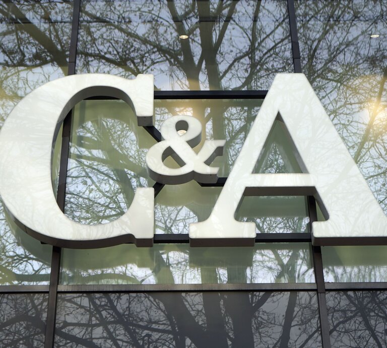 Das Foto zeigt das große weiße Logo "C&A" vor einer Fensterfront, in der sich Bäume spiegeln. Das Logo hebt sich deutlich gegen den durch die Spiegelung erzeugten natürlichen Hintergrund ab. Die Szene wirkt ruhig und die Farben sind harmonisch aufeinander abgestimmt, was einen interessanten visuellen Effekt erzeugt. | © Adobe Stock/ Martin Debus