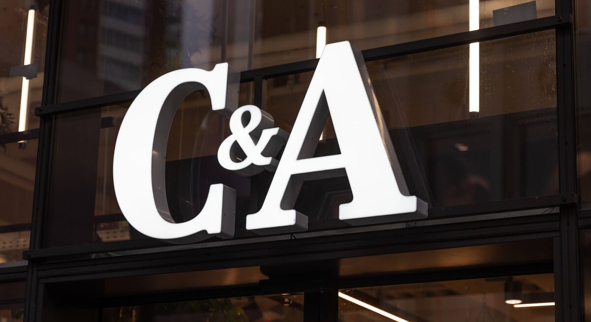  Auf dem Foto ist das beleuchtete Logo des Modeunternehmens C&A zu sehen, das an einem Fenster eines Geschäfts befestigt ist. Die großen, weißen Buchstaben heben sich deutlich gegen die dunkle Umgebung ab. Hinter dem Logo ist eine Innenansicht des Geschäfts mit Pflanzen und einer Reihe von Lichtern zu erkennen, die für eine warme und einladende Atmosphäre sorgen.
