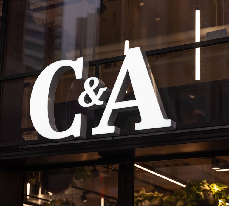  Auf dem Foto ist das beleuchtete Logo des Modeunternehmens C&A zu sehen, das an einem Fenster eines Geschäfts befestigt ist. Die großen, weißen Buchstaben heben sich deutlich gegen die dunkle Umgebung ab. Hinter dem Logo ist eine Innenansicht des Geschäfts mit Pflanzen und einer Reihe von Lichtern zu erkennen, die für eine warme und einladende Atmosphäre sorgen. | © IMAGO / Bihlmayerfotografie