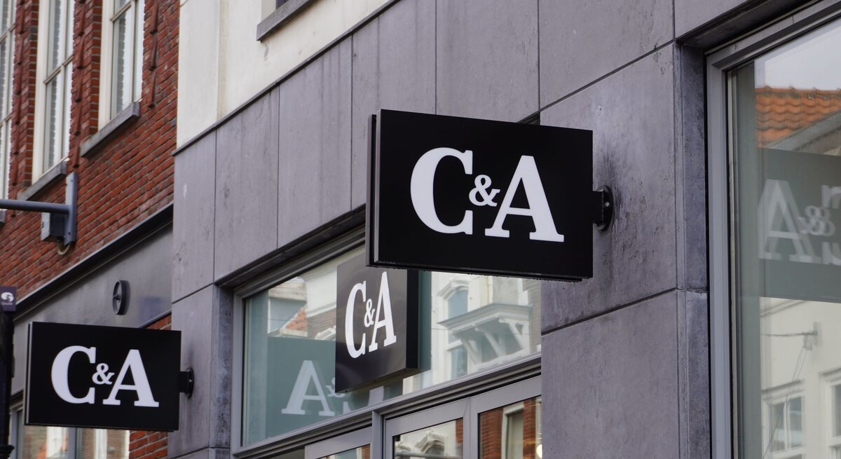 Das Foto zeigt mehrere Schilder des Bekleidungsgeschäfts C&A an der Außenfassade eines modernen Gebäudes. Die Schilder sind schwarz mit dem weißen Logo des Unternehmens darauf. Sie sind so angebracht, dass sie sowohl von Fußgängern auf der Straße als auch von weiter entfernt sichtbar sind. Im Hintergrund sieht man das typische Stadtbild mit Ziegelgebäuden und Fenstern. Das Foto vermittelt einen städtischen Eindruck und hebt die Markenpräsenz im öffentlichen Raum hervor.