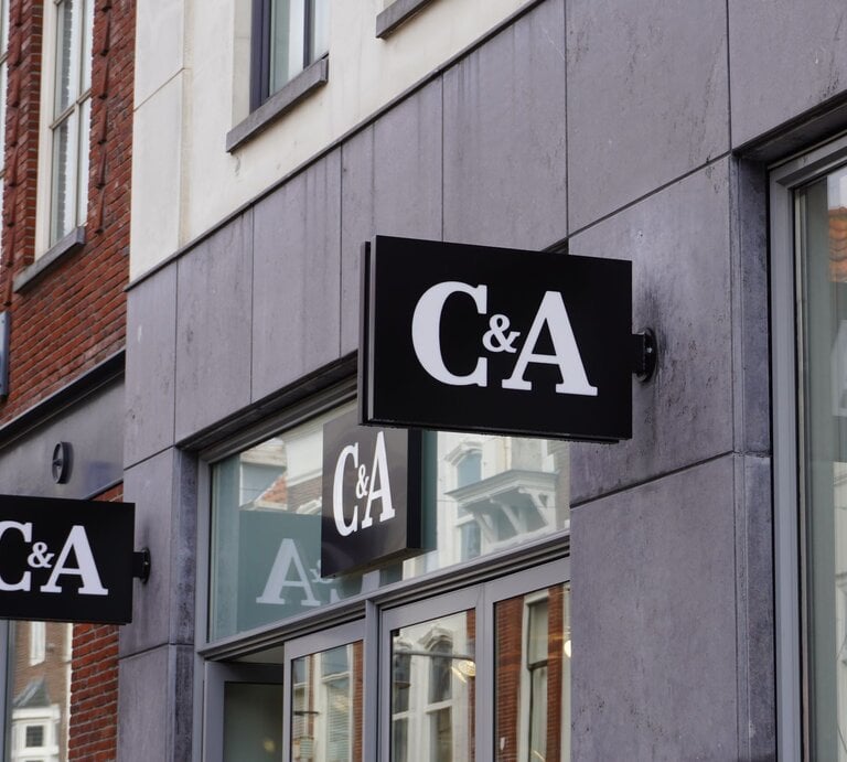 Das Foto zeigt mehrere Schilder des Bekleidungsgeschäfts C&A an der Außenfassade eines modernen Gebäudes. Die Schilder sind schwarz mit dem weißen Logo des Unternehmens darauf. Sie sind so angebracht, dass sie sowohl von Fußgängern auf der Straße als auch von weiter entfernt sichtbar sind. Im Hintergrund sieht man das typische Stadtbild mit Ziegelgebäuden und Fenstern. Das Foto vermittelt einen städtischen Eindruck und hebt die Markenpräsenz im öffentlichen Raum hervor. | © Adobe Stock/ Janet Worg