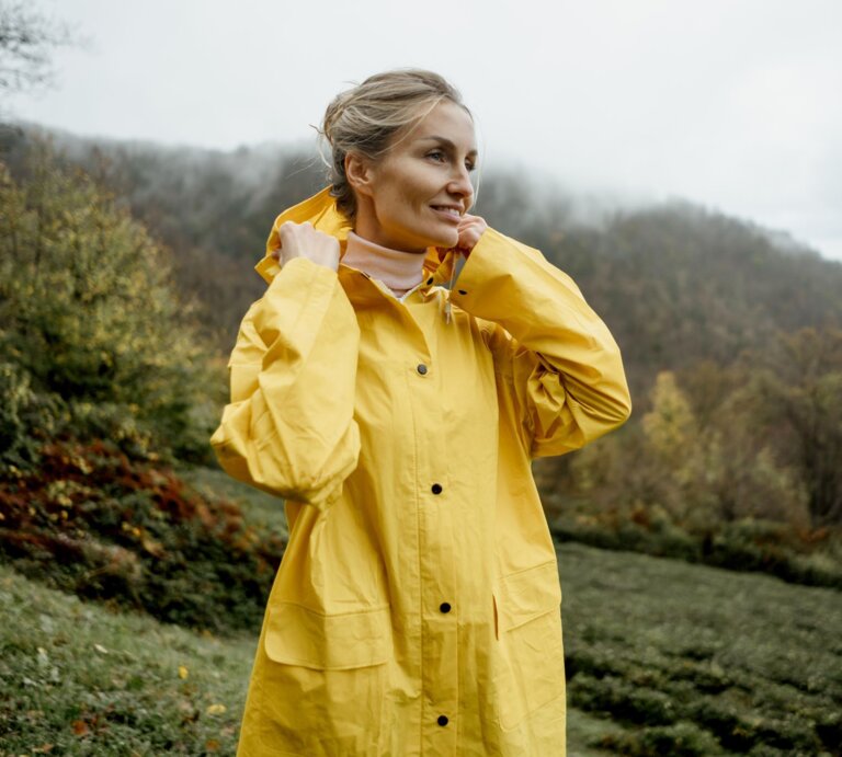 Frau mit hochgesteckten Haaren zieht Kapuze ihres Regenmantels über, vor nebliger Landschaft mit feuchtem Gras und Wald. | © Adobe Stock/ Ilona
