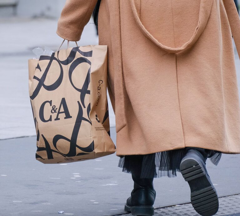 Eine Person geht auf einer Straße und hält eine C&A-Einkaufstüte. Sie trägt einen stilvollen, kamelfarbenen Mantel und schwarze Stiefel, was auf eine kalte Jahreszeit hindeutet. Der Fokus des Fotos liegt auf der Einkaufstüte und der unteren Hälfte der Person, während der obere Teil und der Hintergrund unscharf bleiben. Dies vermittelt das Gefühl des Einkaufens in der Stadt und der Bewegung im Alltagsleben. | © IMAGO / Michael Gstettenbauer