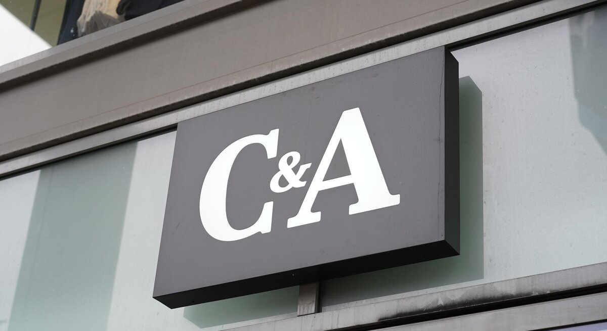 Das Foto zeigt ein Schild des Bekleidungsgeschäfts C&A, angebracht an einer modernen Gebäudefassade. Das Firmenlogo besteht aus großen, weißen Buchstaben auf einem dunklen Hintergrund, was für eine klare und sichtbare Markenidentifikation sorgt. Die Spiegelung im Fenster hinter dem Schild verleiht dem Bild zusätzliche Tiefe und Kontext, indem sie den städtischen Raum widerspiegelt, in dem sich das Geschäft befindet.