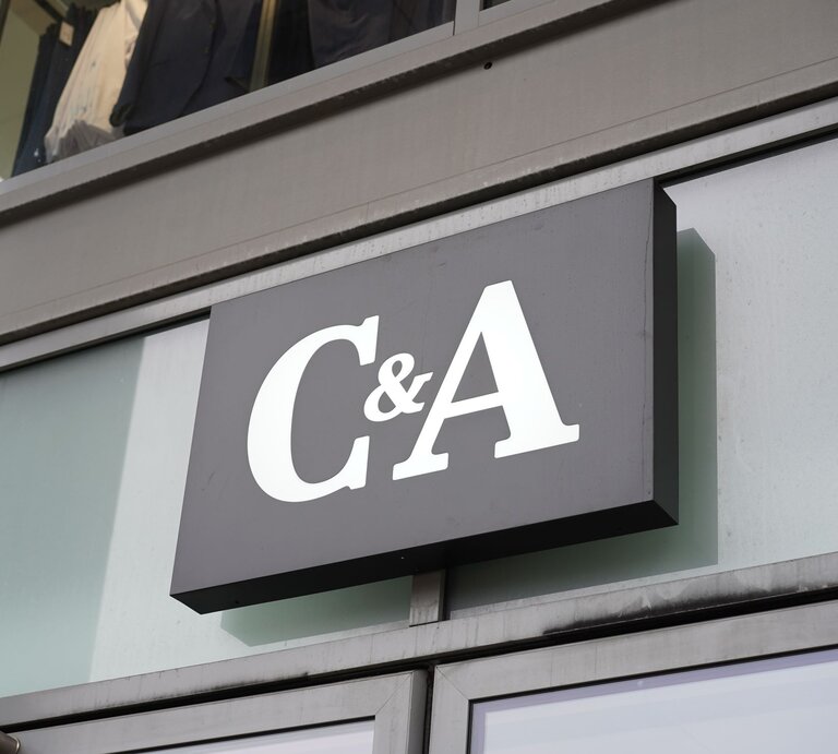 Das Foto zeigt ein Schild des Bekleidungsgeschäfts C&A, angebracht an einer modernen Gebäudefassade. Das Firmenlogo besteht aus großen, weißen Buchstaben auf einem dunklen Hintergrund, was für eine klare und sichtbare Markenidentifikation sorgt. Die Spiegelung im Fenster hinter dem Schild verleiht dem Bild zusätzliche Tiefe und Kontext, indem sie den städtischen Raum widerspiegelt, in dem sich das Geschäft befindet. | © Adobe Stock/ Achim Wagner
