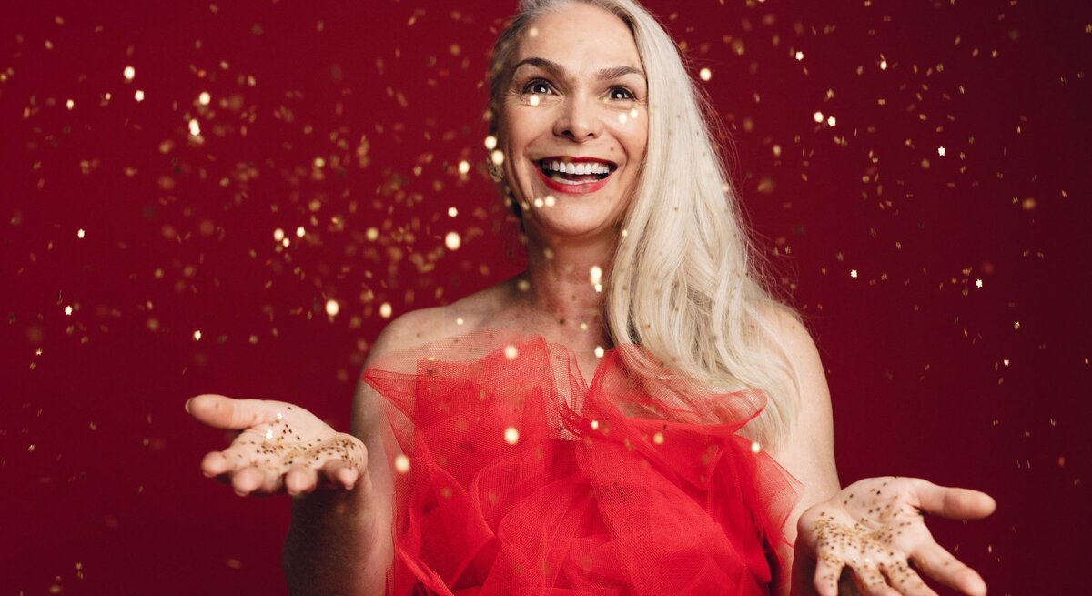 Strahlende Frau mit silbernem Haar in einem roten Kleid wirft goldene Partikel in die Luft, vor leuchtendem roten Hintergrund.