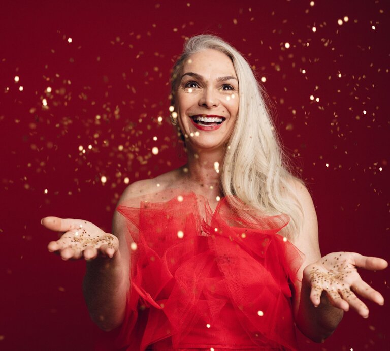 Strahlende Frau mit silbernem Haar in einem roten Kleid wirft goldene Partikel in die Luft, vor leuchtendem roten Hintergrund. | © Adobe Stock/ Jacob Lund
