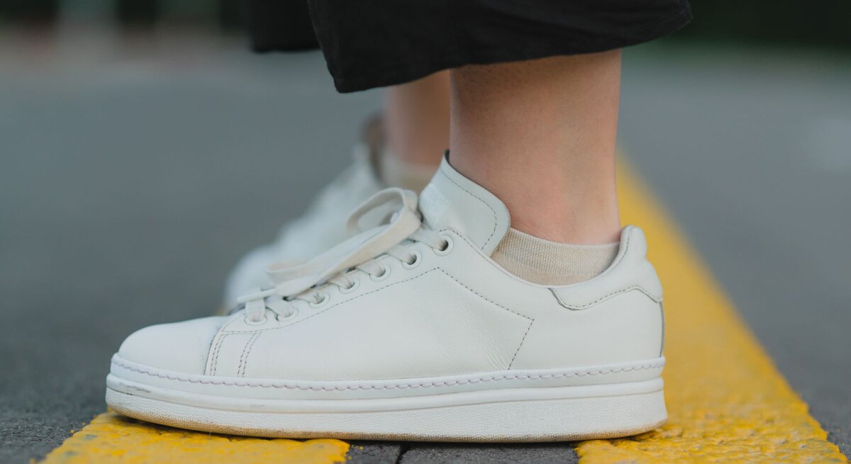 Weiße Sneaker mit beiger Socke auf einer gelben Straßenmarkierung, die von einer Person in schwarzen Hosen getragen werden.