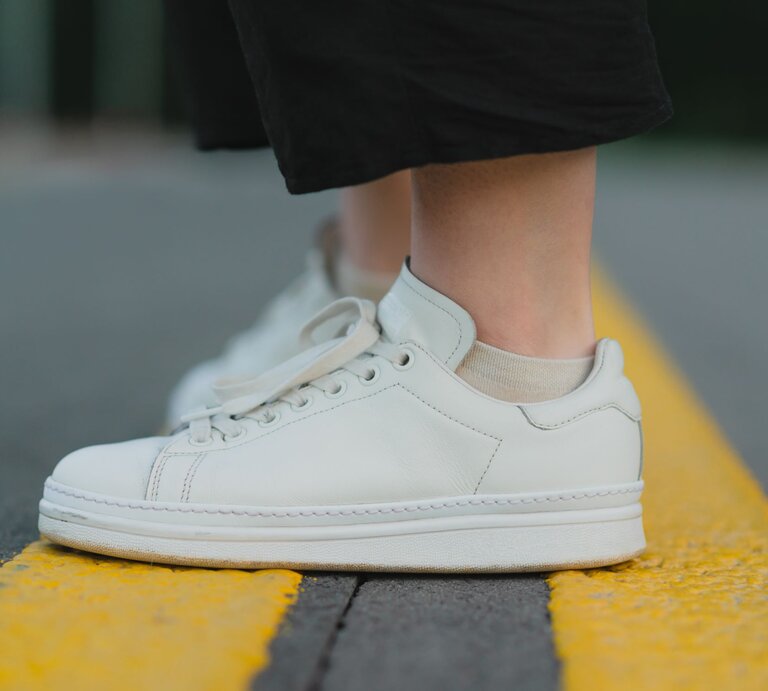 Weiße Sneaker mit beiger Socke auf einer gelben Straßenmarkierung, die von einer Person in schwarzen Hosen getragen werden. | © Adobe Stock/ bodnarphoto
