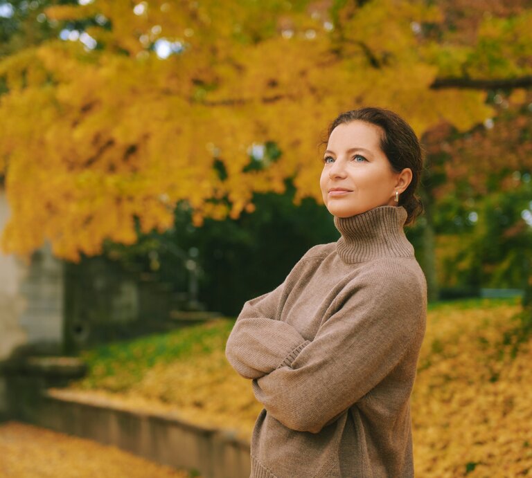 Eine Person in einem hellbraunen Rollkragenpullover steht entspannt vor Herbstbäumen mit goldgelbem Laub und einem steinernen Torbogen. | © Adobe Stock/ annanahabed