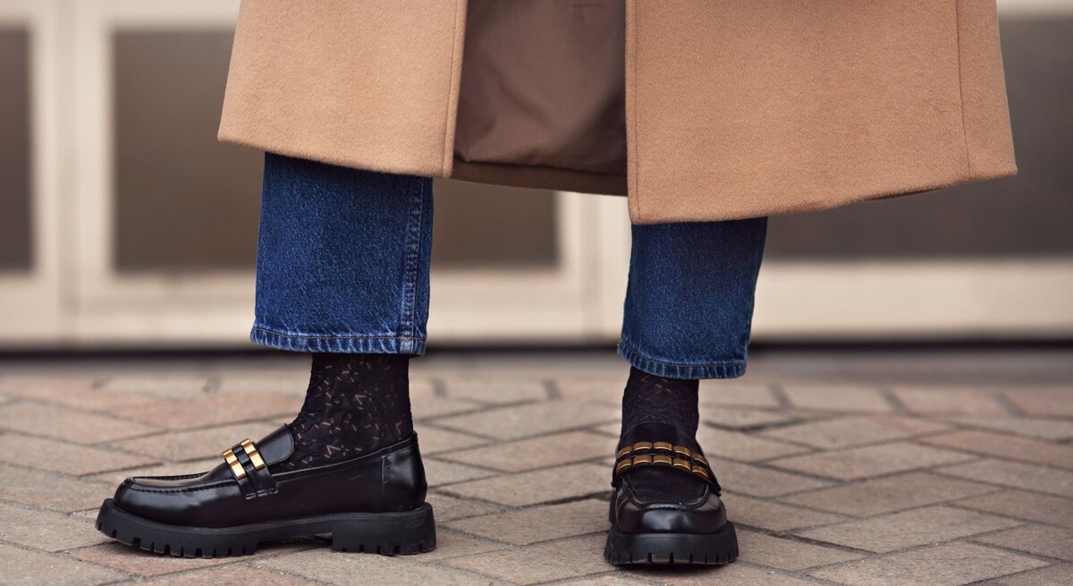 Schwarze Loafer mit goldenen Details und Spitzenstrümpfen, kombiniert mit hochgekrempelten Jeans und einem beigen Mantel auf Pflastersteinen.