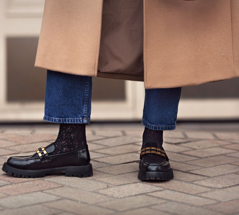 Schwarze Loafer mit goldenen Details und Spitzenstrümpfen, kombiniert mit hochgekrempelten Jeans und einem beigen Mantel auf Pflastersteinen. | © Aobe Stock/ Anna Zhuk