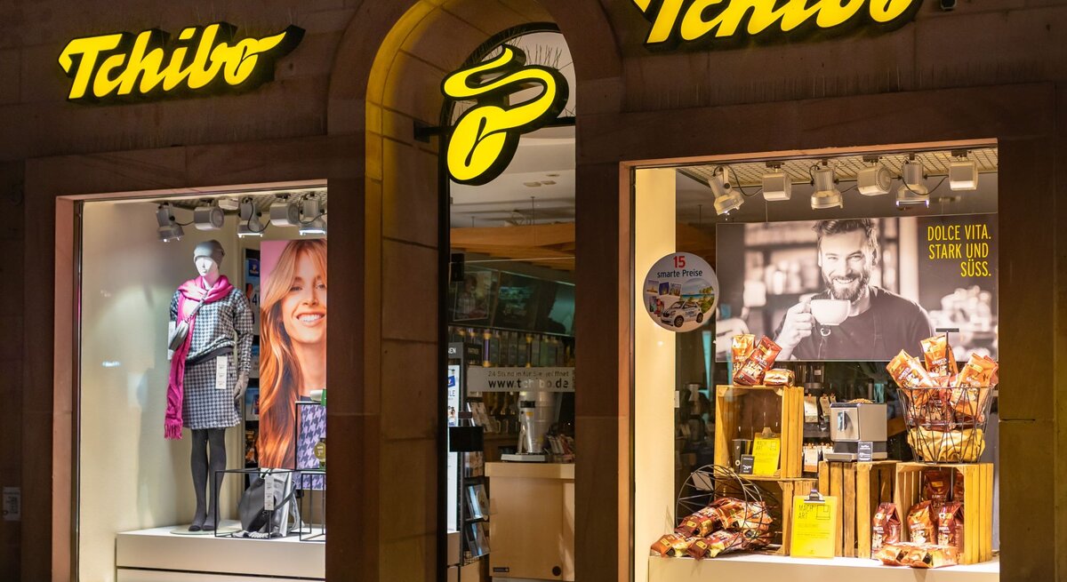 Außenansicht einer Tchibo-Filiale mit gelbem Logo, beleuchtetem Schaufenster, das Kleidung und Kaffeeprodukte präsentiert.