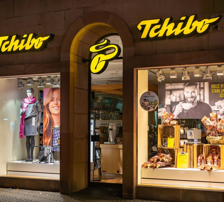 Außenansicht einer Tchibo-Filiale mit gelbem Logo, beleuchtetem Schaufenster, das Kleidung und Kaffeeprodukte präsentiert. | © Adobe Stock/ vulcanus