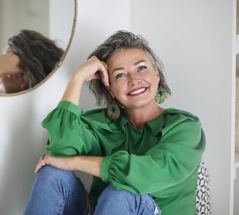 Lachende Frau mittleren Alters mit grauen Haaren, grüner Bluse und Ohrringen lehnt sich entspannt an eine weiße Wand neben einem Spiegel. | © GettyImages/Westend61