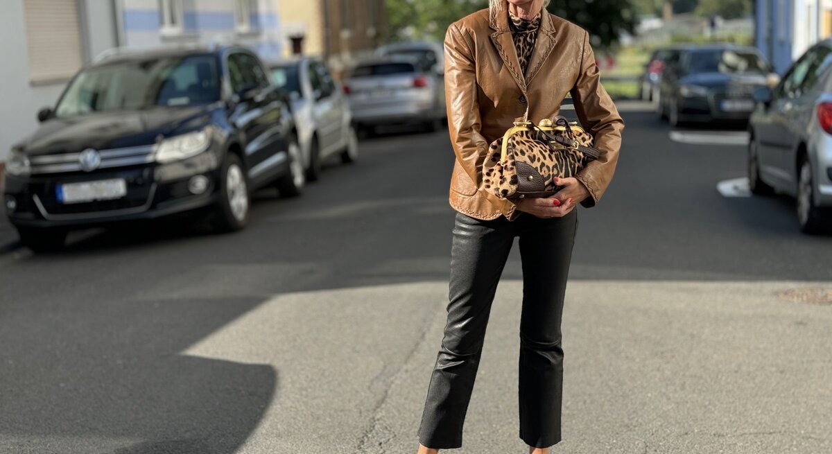 Stilvolle Person, die auf einer Straße steht und eine braune Lederjacke, schwarze, gekürzte Hosen und schwarze Schuhe mit Riemen trägt. Sie hält zudem eine Tasche mit Leopardenmuster und trägt eine Sonnenbrille.