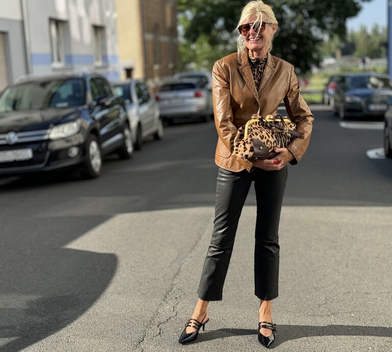 Stilvolle Person, die auf einer Straße steht und eine braune Lederjacke, schwarze, gekürzte Hosen und schwarze Schuhe mit Riemen trägt. Sie hält zudem eine Tasche mit Leopardenmuster und trägt eine Sonnenbrille. | © Renate Zott
