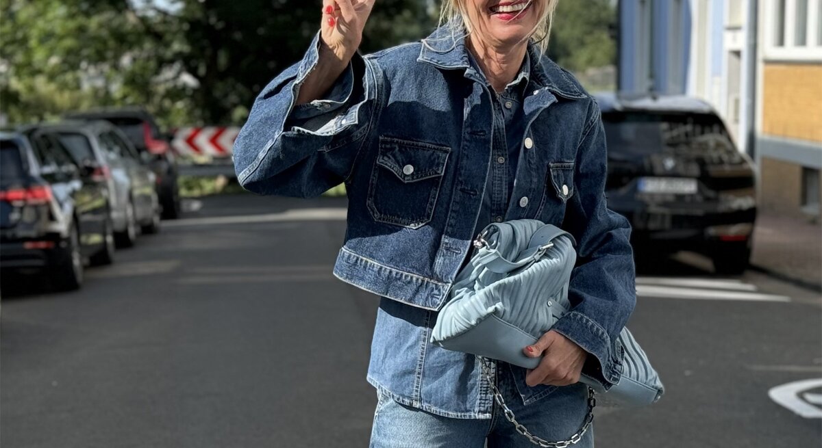 Blonde Frau in einem modischen Jeans-auf-Jeans-Outfit. Sie trägt eine Sonnenbrille und zeigt ein fröhliches Lächeln, während sie ein Peace-Zeichen mit der Hand macht.