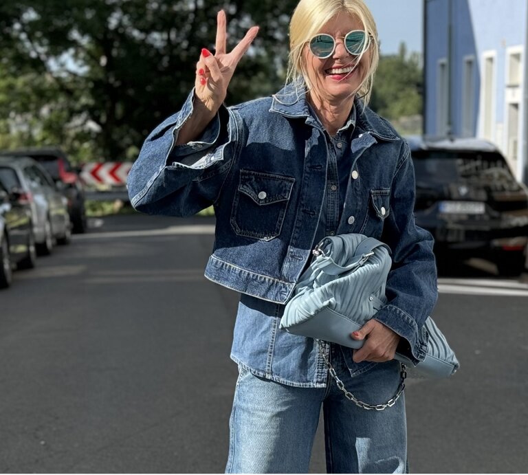 Blonde Frau in einem modischen Jeans-auf-Jeans-Outfit. Sie trägt eine Sonnenbrille und zeigt ein fröhliches Lächeln, während sie ein Peace-Zeichen mit der Hand macht. | © Renate Zott
