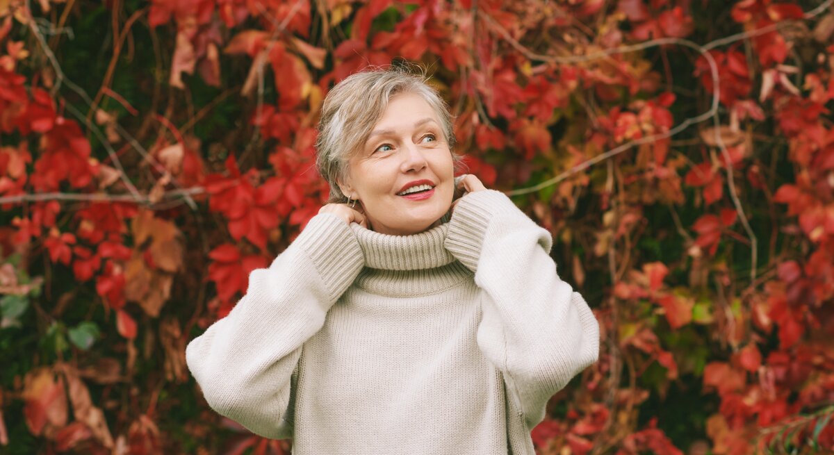 Eine Person in cremeweißem Rollkragenpullover steht vor einer herbstlichen Wand aus roten und grünen Blättern.