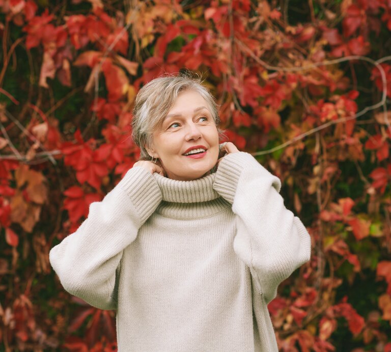 Eine Person in cremeweißem Rollkragenpullover steht vor einer herbstlichen Wand aus roten und grünen Blättern. | © Adobe Stock/ annanahabed