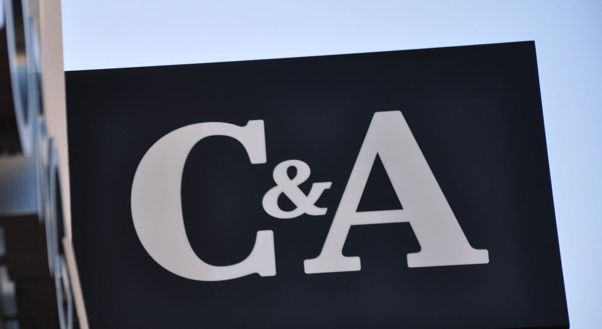  Ein großes, schlichtes Schild mit den Buchstaben "C&A" in weißer Farbe auf einem schwarzen Hintergrund. Das Schild ist schräg fotografiert gegen einen klaren blauen Himmel. Die Sonne scheint von der Seite und beleuchtet das Schild und seine Kanten.