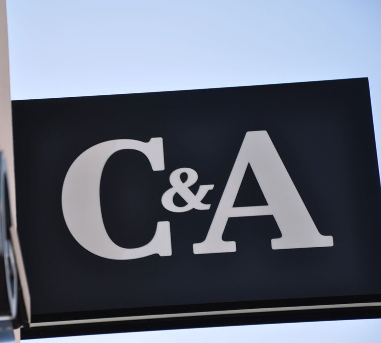  Ein großes, schlichtes Schild mit den Buchstaben "C&A" in weißer Farbe auf einem schwarzen Hintergrund. Das Schild ist schräg fotografiert gegen einen klaren blauen Himmel. Die Sonne scheint von der Seite und beleuchtet das Schild und seine Kanten. | © Adobe Stock/ CSschmuck