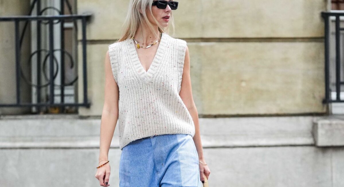 Junge blonde Frau in beigefarbenem Stricktop und weiter Patchwork-Jeans geht mit kleiner Tasche auf herbstlicher Straße.