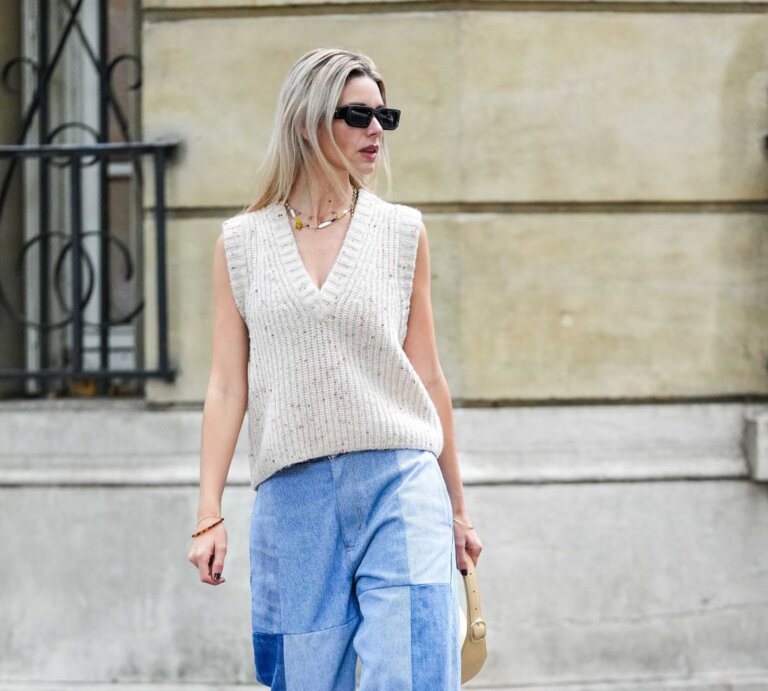 Junge blonde Frau in beigefarbenem Stricktop und weiter Patchwork-Jeans geht mit kleiner Tasche auf herbstlicher Straße. | © Edward Berthelot/Getty Images