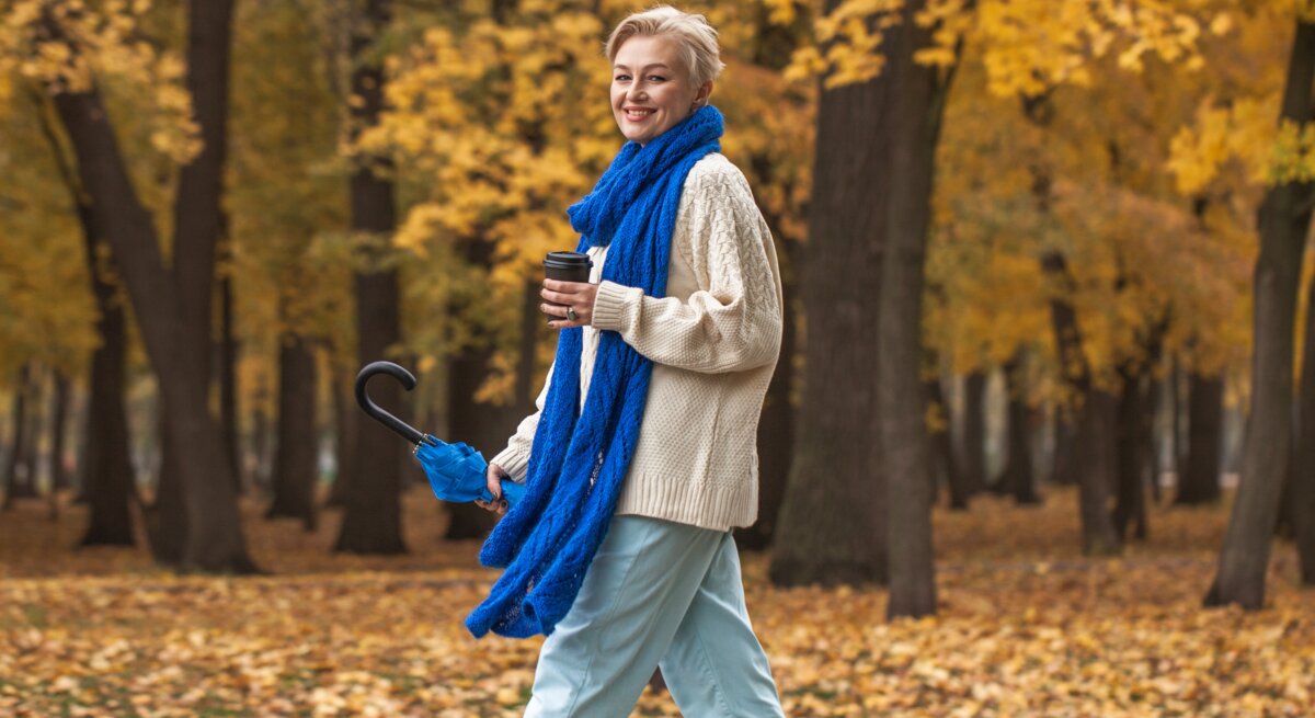 Eine Person im cremefarbenen Pullover und blauer Jeans spaziert mit Regenschirm und Kaffee durch einen herbstlichen Park voller Laub.