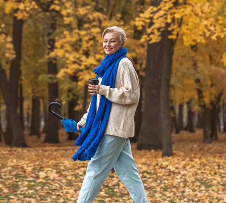 Eine Person im cremefarbenen Pullover und blauer Jeans spaziert mit Regenschirm und Kaffee durch einen herbstlichen Park voller Laub. | © Adobe Stock/ Andrey_Arkusha