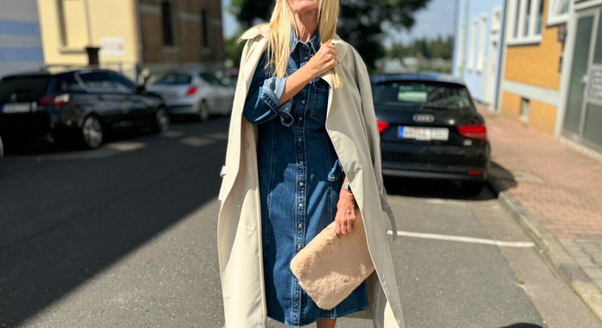 Frau, die ein stilvolles Jeanskleid trägt, das sie mit einem beigen Mantel über den Schultern kombiniert. Sie hält eine flauschige Tasche in der Hand und trägt beigefarbene Stiefeletten.