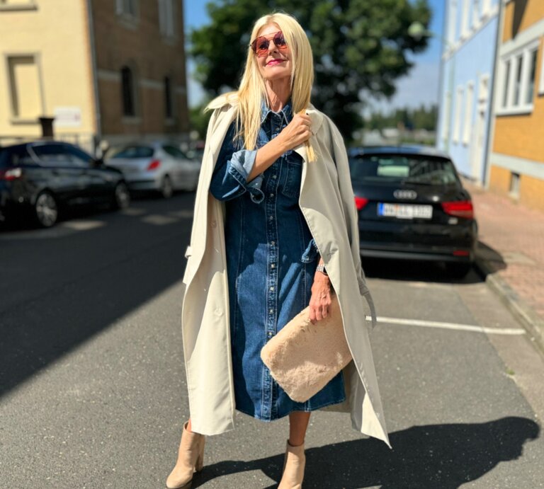 Frau, die ein stilvolles Jeanskleid trägt, das sie mit einem beigen Mantel über den Schultern kombiniert. Sie hält eine flauschige Tasche in der Hand und trägt beigefarbene Stiefeletten. | © Renate Zott