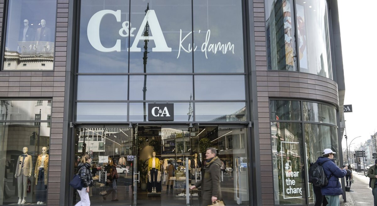 Auf diesem Foto ist eine Filiale von C&A zu sehen, welche an einem offensichtlich urbanen und belebten Standort liegt. Auffällig ist die große Beschriftung "C&A Ku'damm" auf den Fenstern, was darauf hindeutet, dass sich das Geschäft am oder in der Nähe des bekannten Kurfürstendamms in Berlin befinden könnte. Die großflächigen Schaufenster präsentieren verschiedene Modelle, die aktuelle Modekollektionen darstellen. Vorbeigehende Passanten vermitteln eine Atmosphäre der Geschäftigkeit, was typisch für eine Einkaufsstraße in einer Großstadt ist. Das Foto fängt das dynamische Treiben des städtischen Alltags ein und zeigt gleichzeitig die Markenpräsenz von C&A im Einzelhandel.