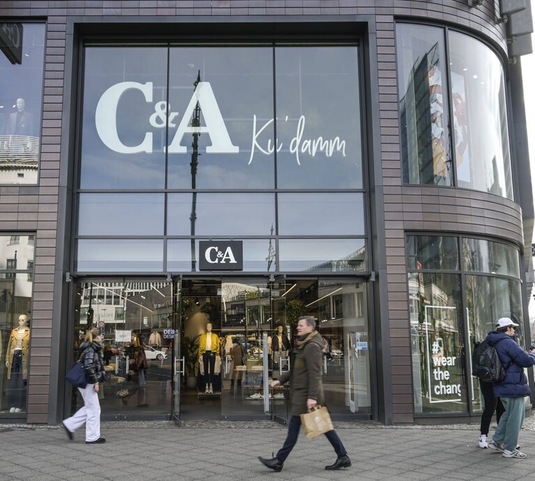 Auf diesem Foto ist eine Filiale von C&A zu sehen, welche an einem offensichtlich urbanen und belebten Standort liegt. Auffällig ist die große Beschriftung "C&A Ku'damm" auf den Fenstern, was darauf hindeutet, dass sich das Geschäft am oder in der Nähe des bekannten Kurfürstendamms in Berlin befinden könnte. Die großflächigen Schaufenster präsentieren verschiedene Modelle, die aktuelle Modekollektionen darstellen. Vorbeigehende Passanten vermitteln eine Atmosphäre der Geschäftigkeit, was typisch für eine Einkaufsstraße in einer Großstadt ist. Das Foto fängt das dynamische Treiben des städtischen Alltags ein und zeigt gleichzeitig die Markenpräsenz von C&A im Einzelhandel. | © IMAGO / Schöning