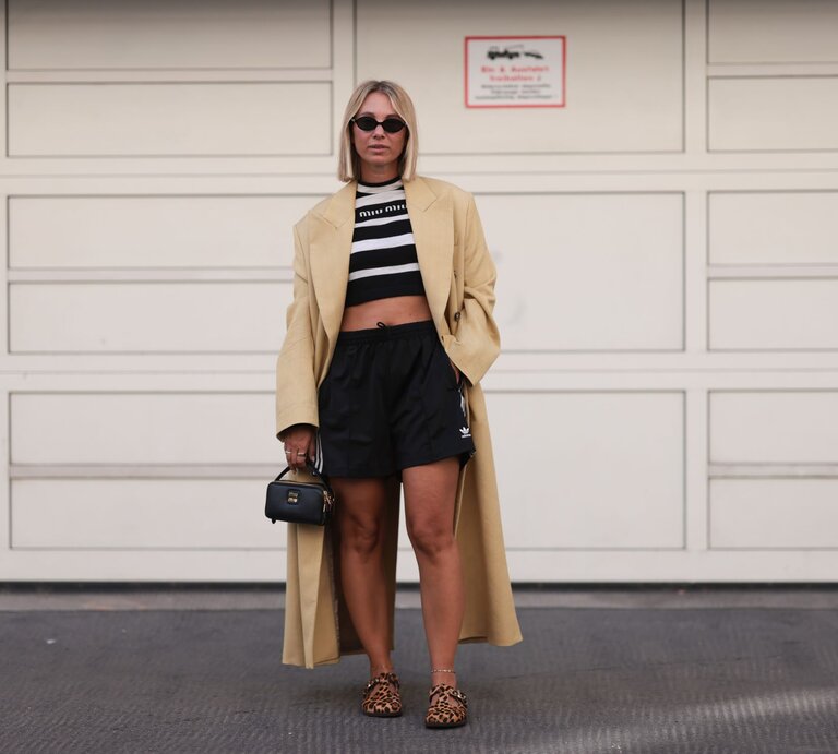 Modisch gekleidete Frau trägt langen beigen Mantel, schwarze Shorts, gestreiftes Top und Leoparden-Slipper, stehend auf grauem Asphalt. | © Jeremy Moeller/Getty Images