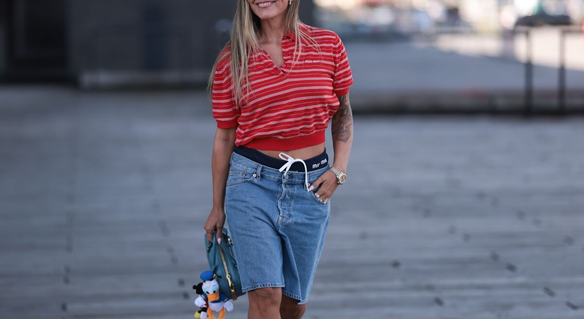 Lächelnde Frau in sommerlichem Outfit mit Jeansshorts, rot gestreiftem Shirt und Tätowierung am rechten Arm, hält eine blaue Tasche mit Stoffanhängern.
