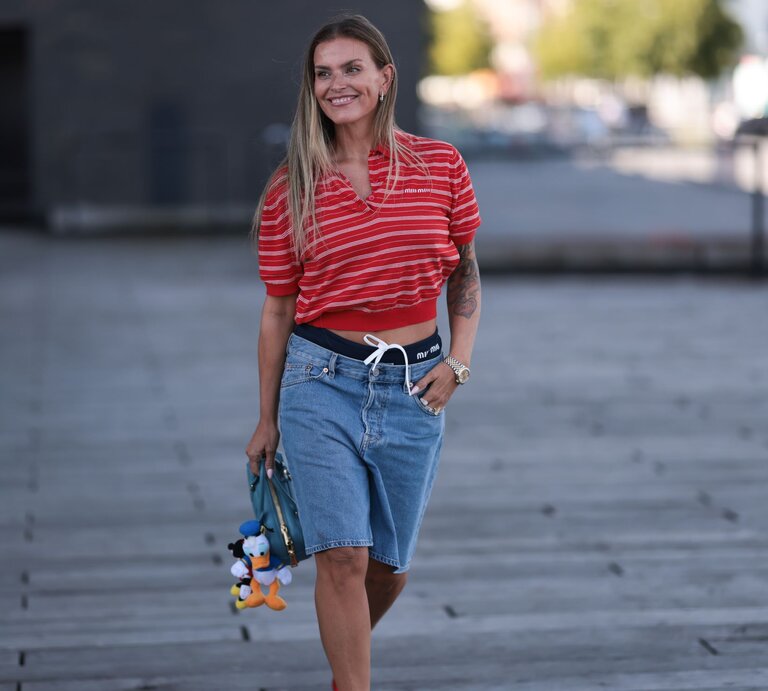 Lächelnde Frau in sommerlichem Outfit mit Jeansshorts, rot gestreiftem Shirt und Tätowierung am rechten Arm, hält eine blaue Tasche mit Stoffanhängern. | © Jeremy Moeller/Getty Images