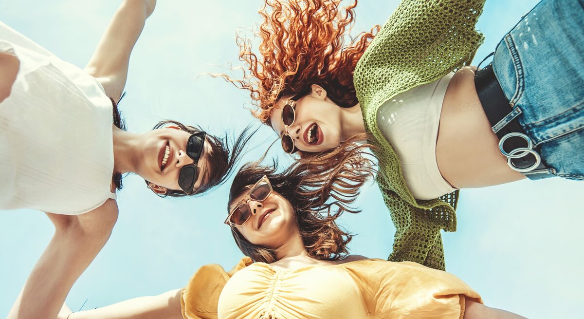Lächelnde Frauen in sommerlicher Kleidung und Sonnenbrillen, von unten fotografiert, mit ausgestreckten Armen unter blauem Himmel.