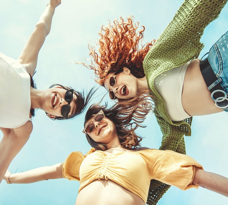 Lächelnde Frauen in sommerlicher Kleidung und Sonnenbrillen, von unten fotografiert, mit ausgestreckten Armen unter blauem Himmel. | © Getty Images/ GizemBDR