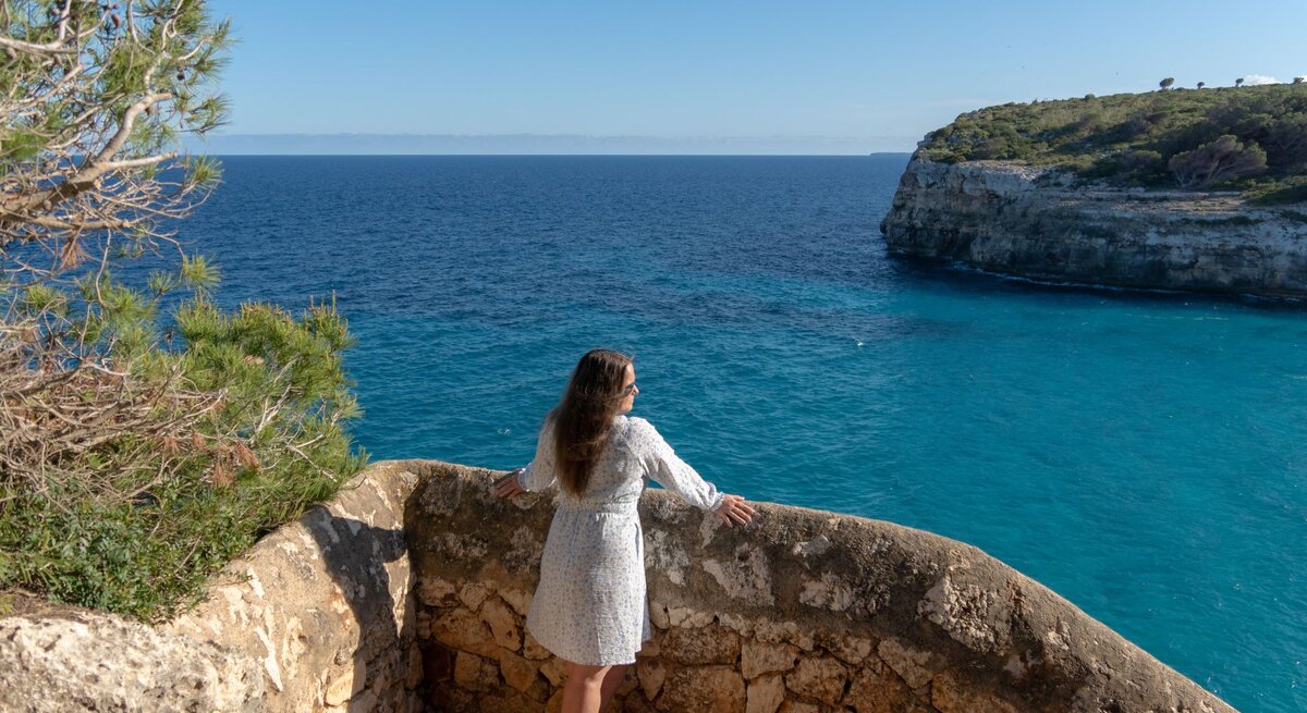 Frau in hellem Sommerkleid und Sandalen blickt entspannt von steinerner Aussichtsplattform auf türkisblaues Meer und steile Küste auf Mallorca.