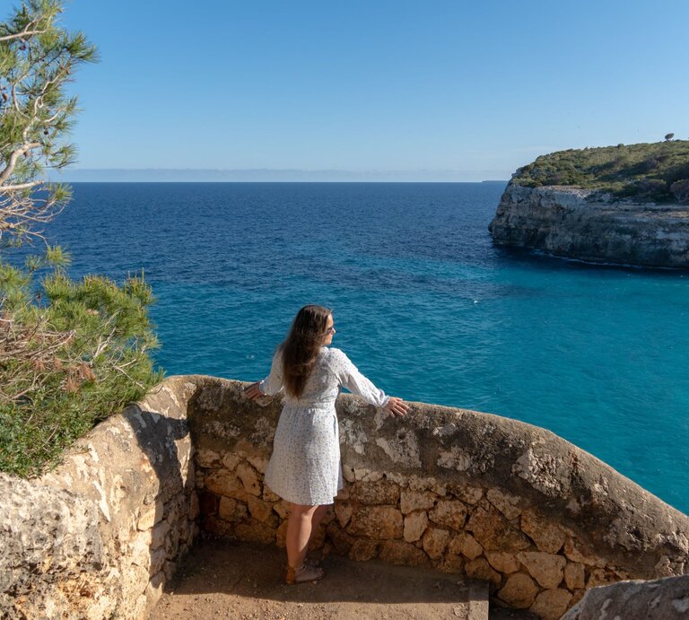 Frau in hellem Sommerkleid und Sandalen blickt entspannt von steinerner Aussichtsplattform auf türkisblaues Meer und steile Küste auf Mallorca. | © Adobe Stock/ Marc Kunze