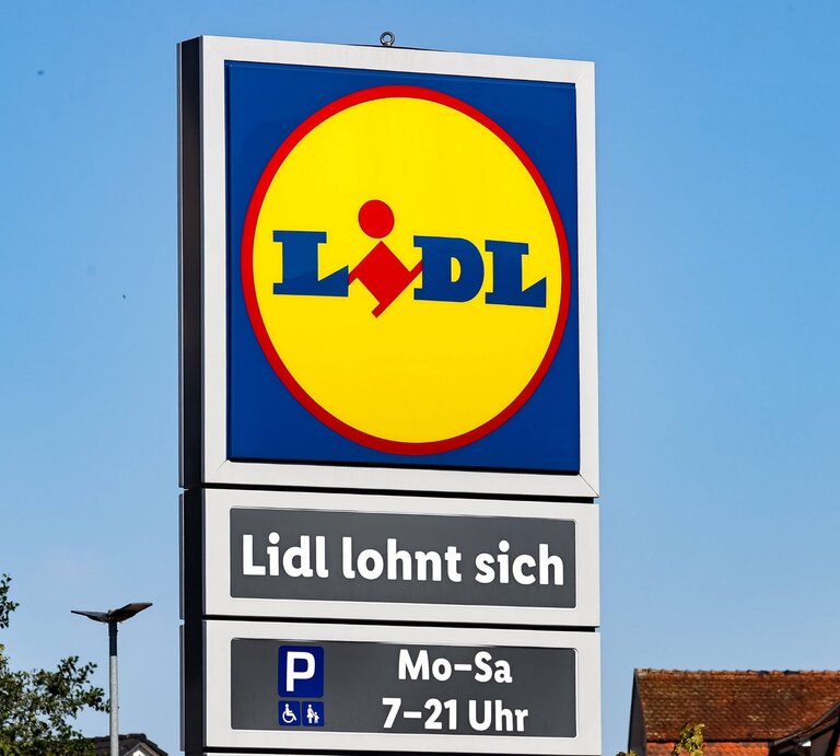 Ein Schild mit dem Lidl-Logo und dem Slogan "Lidl lohnt sich" vor klarem, blauem Himmel und Gebäuden im Hintergrund. | © Imago Images/ xGrantxHubbsx