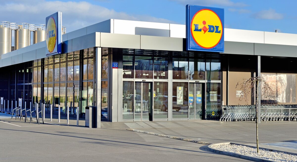 Auf dem Bild ist eine moderne Lidl-Filiale zu sehen. Sie hat eine schlichte und zeitgenössische Architektur mit einer klaren Glasfront und einem flachen Dach. Über dem Eingang ist ein großes, beleuchtetes Lidl-Logo angebracht. Vor dem Supermarkt befinden sich ordentlich aufgereihte Einkaufswagen. Im Hintergrund sind silberfarbene Silos zu sehen, was auf eine industrielle Umgebung hindeutet. Die Szene ist bei Tageslicht aufgenommen, mit einem klaren blauen Himmel im Hintergrund.