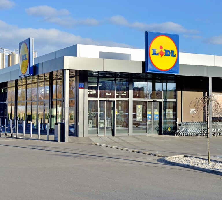 Auf dem Bild ist eine moderne Lidl-Filiale zu sehen. Sie hat eine schlichte und zeitgenössische Architektur mit einer klaren Glasfront und einem flachen Dach. Über dem Eingang ist ein großes, beleuchtetes Lidl-Logo angebracht. Vor dem Supermarkt befinden sich ordentlich aufgereihte Einkaufswagen. Im Hintergrund sind silberfarbene Silos zu sehen, was auf eine industrielle Umgebung hindeutet. Die Szene ist bei Tageslicht aufgenommen, mit einem klaren blauen Himmel im Hintergrund. | © Adobe Stock/ Tomasz Warszewski