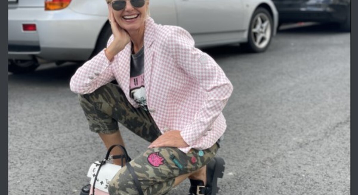 Lächelnde Frau mittleren Alters mit blonden Haaren und Sonnenbrille, die in einer Hockstellung auf einem Parkplatz posiert. Sie trägt eine rosafarbene, karierte Jacke, ein schwarzes T-Shirt mit Aufdruck, Tarnhosen und schwarze Schuhe.
