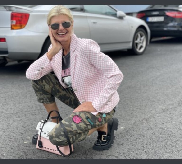 Lächelnde Frau mittleren Alters mit blonden Haaren und Sonnenbrille, die in einer Hockstellung auf einem Parkplatz posiert. Sie trägt eine rosafarbene, karierte Jacke, ein schwarzes T-Shirt mit Aufdruck, Tarnhosen und schwarze Schuhe. | © Renate Zott