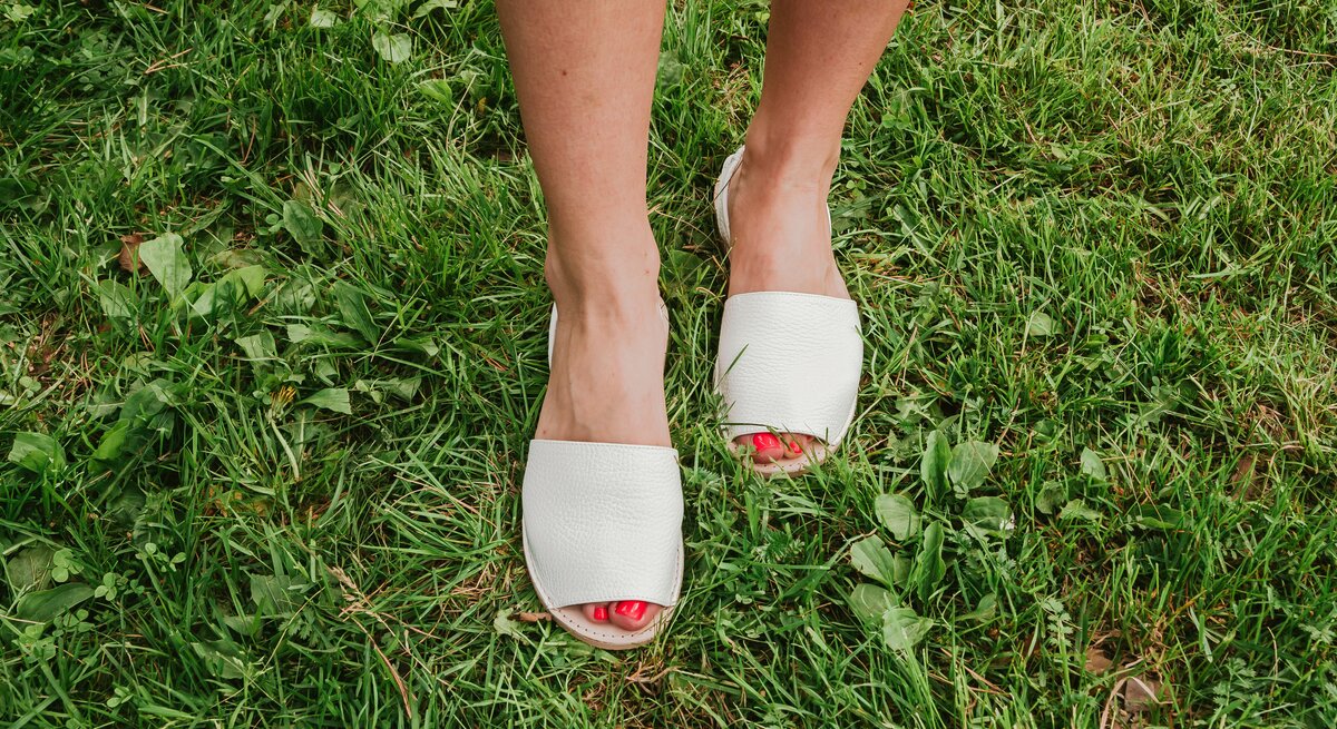 Weibliche Füße mit rotem Nagellack in weißen Espadrilles-Sandalen stehen auf frisch gemähtem, saftig grünem Rasen.