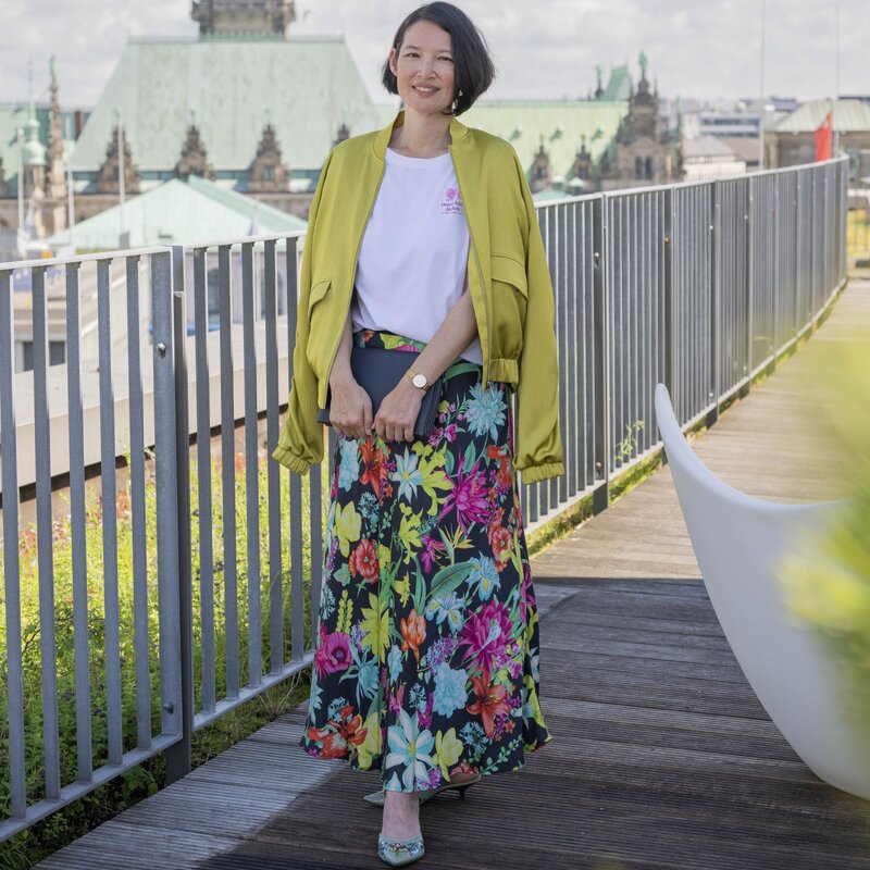 Frau mit kurzen schwarzen Haaren, gelber Jacke, weißem T-Shirt und buntem Blumenrock steht lächelnd auf einer Dachterrasse vor dem Hamburger Rathaus. | © Bild der Frau/ Ulrike Schacht