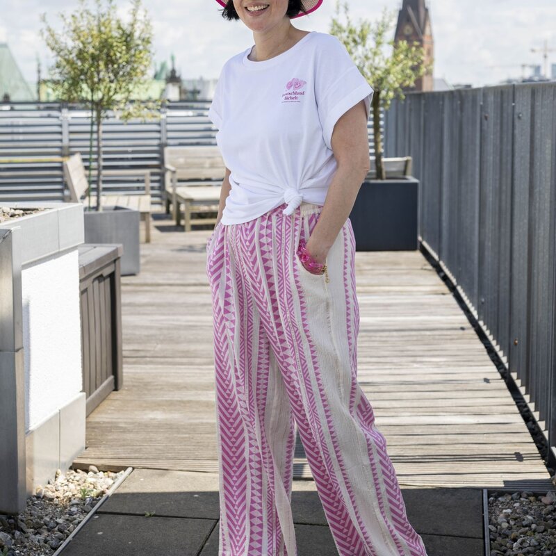 Frau mit Strohhut, rosa Sonnenbrille, weißem T-Shirt und rosa-weiß gemusterten Hosen steht lächelnd auf einer Dachterrasse. | © Bild der Frau/ Ulrike Schacht